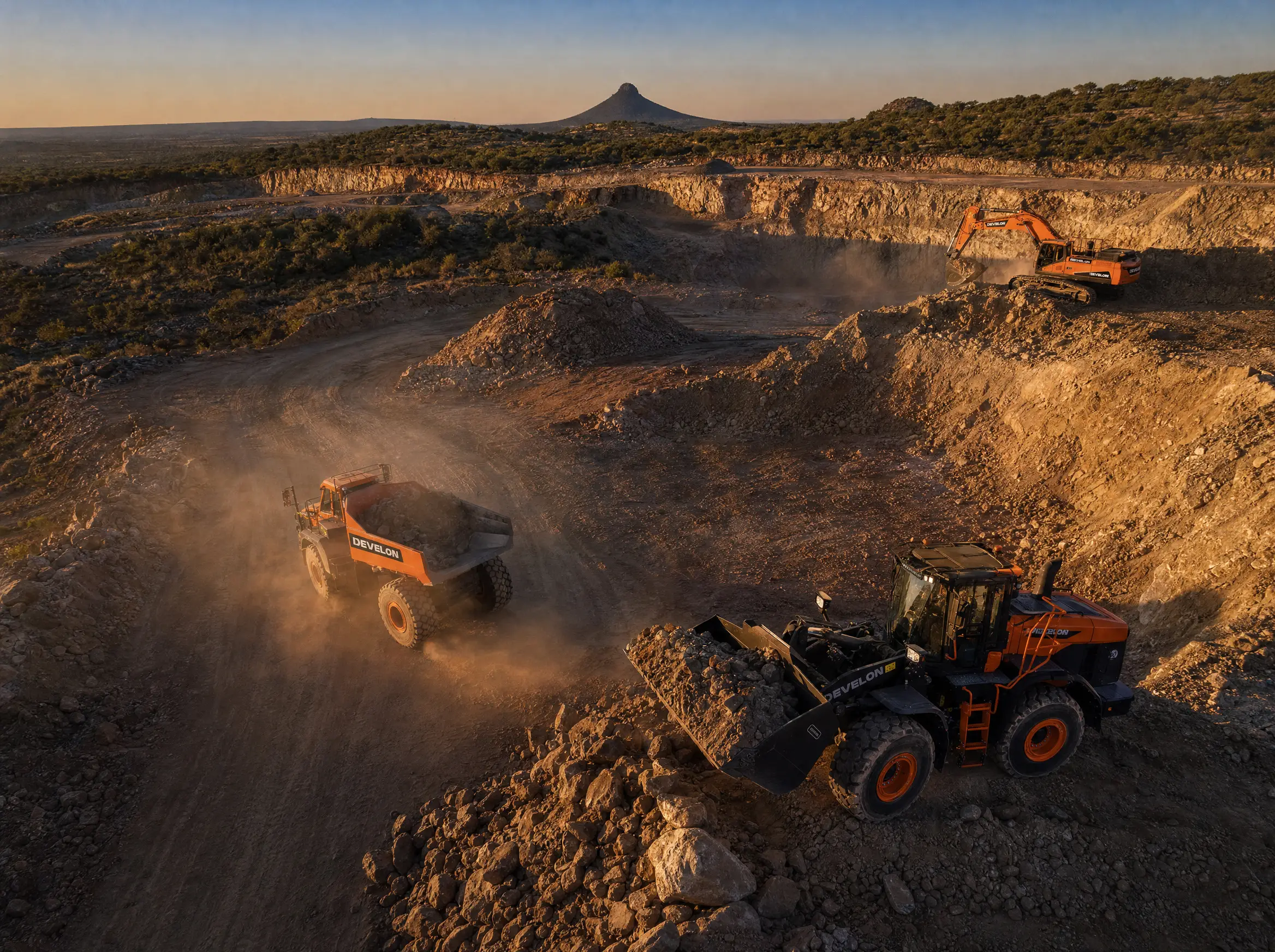 Oblique aerial of a working mine site: a loaded DEVELON dump truck, a wheel loader and an excavator, a koppie on the horizon